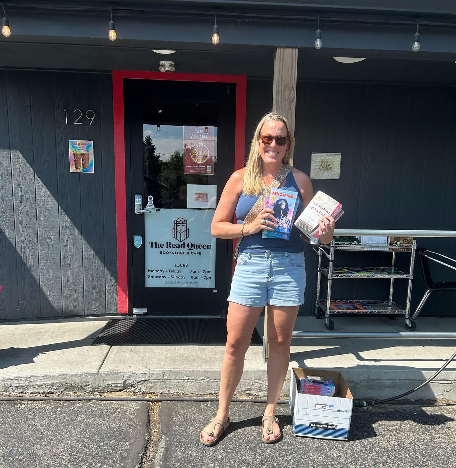 Jamie Dickinson outside The Read Queen bookstore and cafe, holding two hardbacks, a filing box of books at her feet.